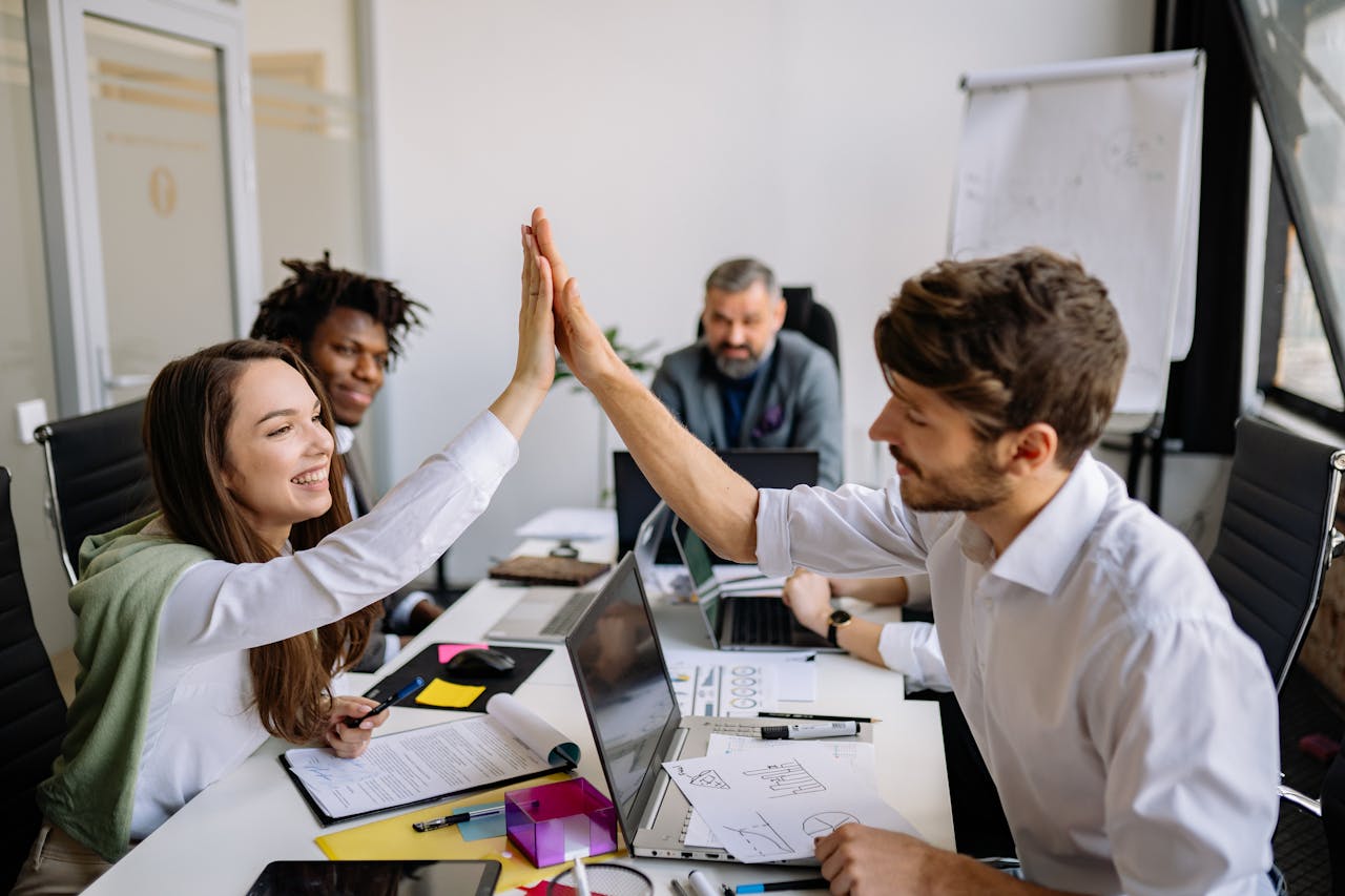 A diverse group of colleagues in an office, celebrating with a high five during a meeting, showcasing teamwork and collaboration.